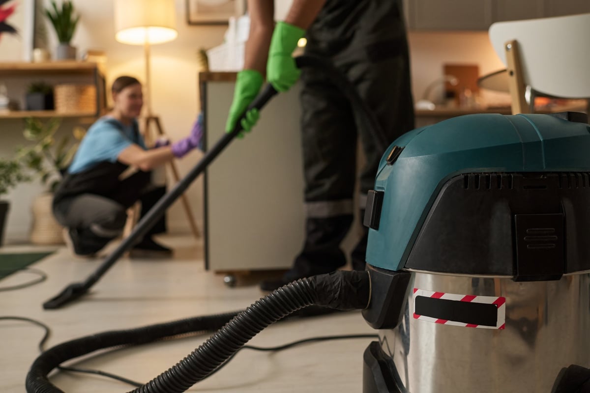 Close-up of worker using vacuum cleaner during housework with his colleague in apartment